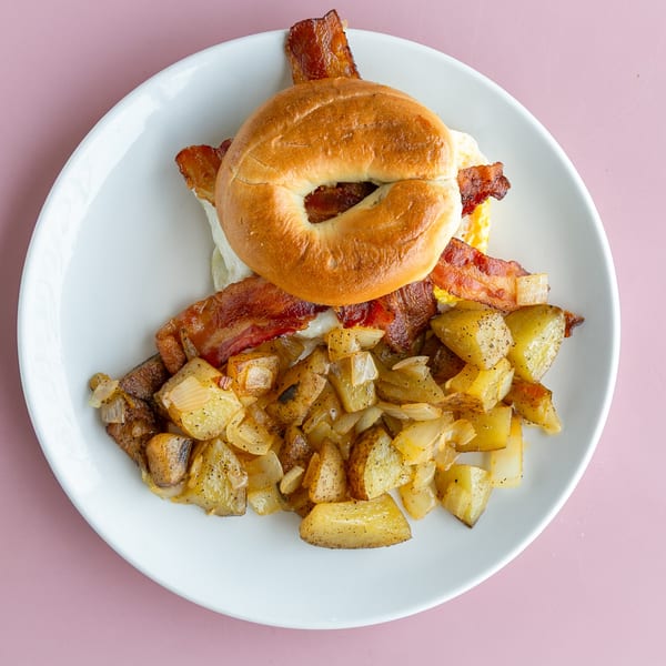 Breakfast plate with a bagel sandwich, bacon, scrambled eggs, and home fries with onions.