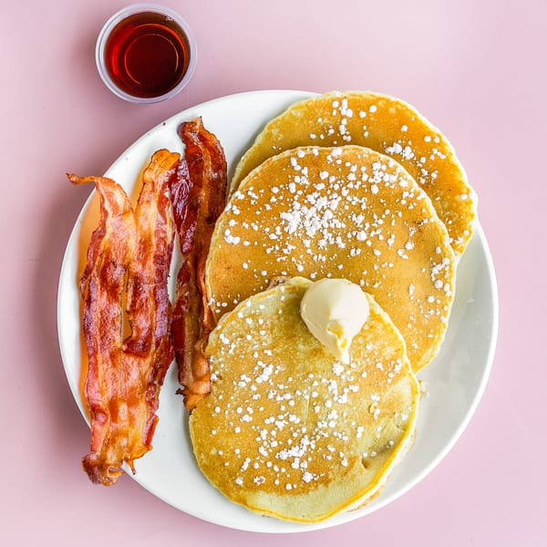 A plate of pancakes with powdered sugar and butter, crispy bacon, and a side of syrup.