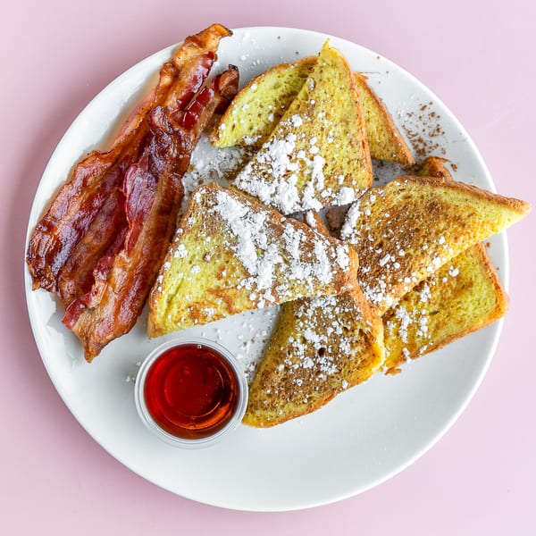 A white plate with French toast, bacon, and a side of syrup on a pink background.