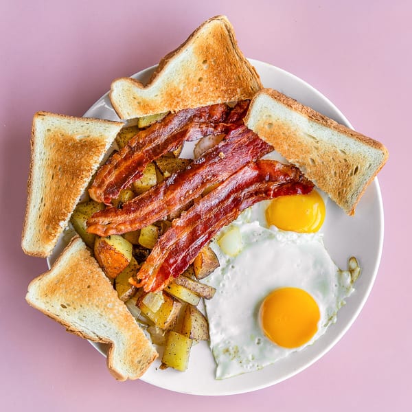 Breakfast plate with crispy bacon, fried eggs, home fries, and toast on a pink background.